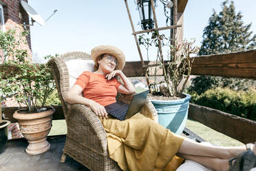Porträt einer nachdenklichen reifen Frau mit Laptop, die sich auf einer Terrasse entspannt - KMKF01315