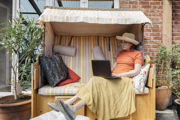 Ältere Frau sitzt auf der Terrasse und arbeitet am Laptop - KMKF01318