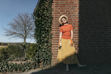 Portrait of mature woman with gardening gloves and pruner standing in front of brick wall enjoying sunlight - KMKF01330