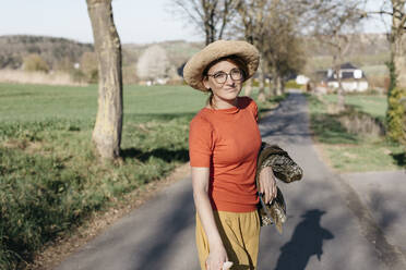 Porträt einer lächelnden reifen Frau mit Brille und Strohhut, die auf einer Landstraße steht - KMKF01337