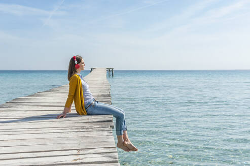 Frau sitzt auf einem Bootssteg und hört mit Kopfhörern Musik, Mallorca, Spanien - DIGF10339