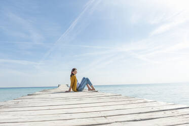Frau sitzt auf einem Bootssteg und hört mit Kopfhörern Musik, Mallorca, Spanien - DIGF10341