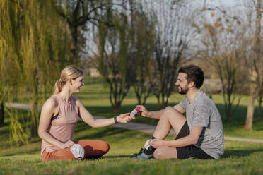 Ein lächelnder Mann gibt einer Frau Handdesinfektionsmittel, während er im Park auf dem Rasen sitzt - STDF00226