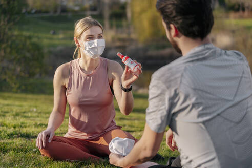 Frau mit Gesichtsmaske zeigt Mann Handdesinfektionsmittel, während er im Park sitzt - STDF00227