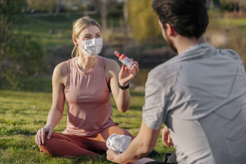 Woman wearing face mask showing hand sanitizer to man while sitting at park - STDF00227