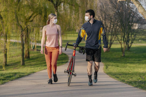 Young couple walking with bicycle on footpath at park during coronavirus pandemic - STDF00228