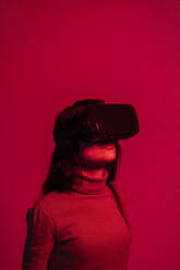 Mid adult woman using virtual reality headset while standing against red background - EGAF00005