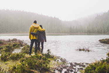 Full length rear view of young couple standing while looking at river near forest during rainy season - FVSF00232