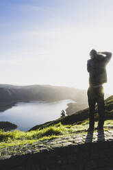 Rear view of man photographing Lagoa do Fogo against sky during sunset, San Miguel, Azores, Portugal - FVSF00246