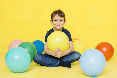 Porträt von lächelnden niedlichen Jungen sitzen mit bunten Luftballons gegen gelben Hintergrund - JRFF04419