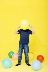 Boy holding yellow balloon in front of face while standing against colored background - JRFF04421