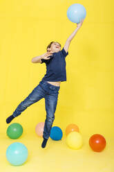 Full length of boy catching balloon while playing against yellow background - JRFF04422