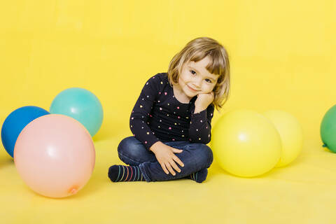 Porträt des lächelnden niedlichen Mädchen sitzt mit bunten Luftballons gegen gelben Hintergrund, lizenzfreies Stockfoto