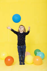 Cute girl catching balloon while standing against yellow background - JRFF04424