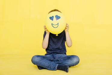 Boy holding yellow balloon with anthropomorphic face against colored background - JRFF04429