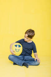 Full length of cute boy looking at yellow balloon with anthropomorphic face while sitting against colored background - JRFF04430