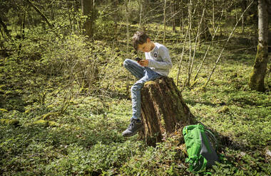 Full length of boy sitting on tree stump while using smart phone in Swabian Jura forest during hiking - DIKF00476