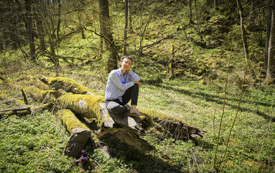Entspannte Frau in voller Länge auf moosbewachsenem Baumstamm in einem Wald auf der Schwäbischen Alb sitzend - DIKF00478