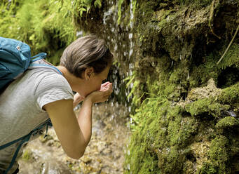 Woman drinking water from her hands cupped under trickle of Urach Waterfall in forest - DIKF00494