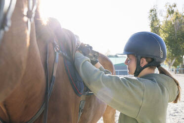 Seitenansicht eines Teenagers, der seinen Sattel vor dem Reiten auf dem Trainingsplatz einstellt - ABZF03103