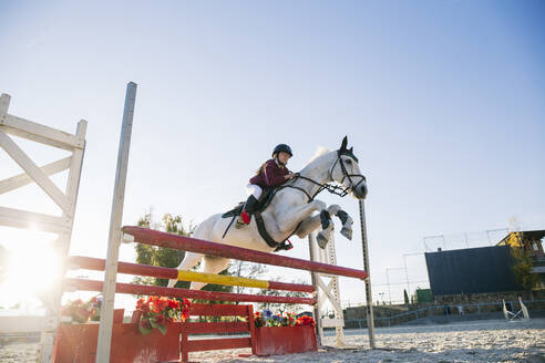 Low angle view of teenage jockey girl riding white horse over hurdle on training ground against clear sky - ABZF03114