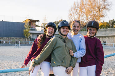 Porträt von fröhlichen weiblichen Jockeys, die auf dem Trainingsplatz gegen den Himmel zusammenstehen - ABZF03119