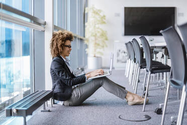 Smiling young businesswoman sitting on the floor in conference room using laptop - DIGF10397