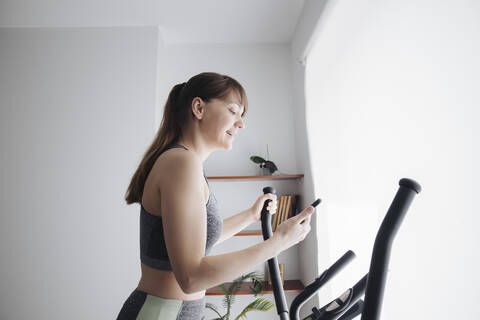 Woman using smartphone while performing workout on elliptical trainer at home stock photo