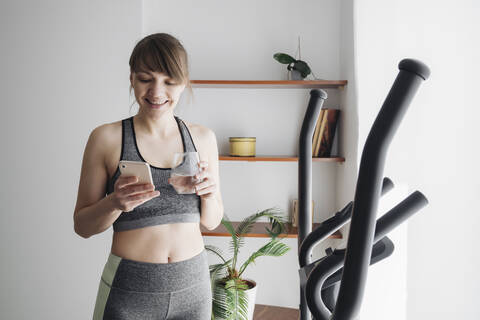 Frau, die Wasser trinkt und ihr Smartphone benutzt, nachdem sie zu Hause auf dem Ellipsentrainer trainiert hat, lizenzfreies Stockfoto