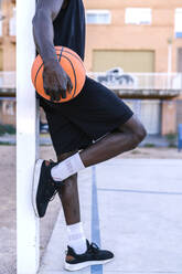 Young man holding basketball leaning on pole on basketball court - EGAF00021