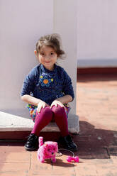 Porträt eines lächelnden kleinen Mädchens, das mit seinem rosa Spielzeughund auf einem Dach sitzt - EGAF00037