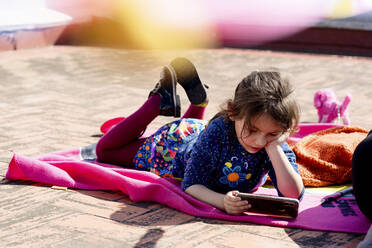 Porträt eines kleinen Mädchens, das auf einer Decke auf dem Dach liegt und auf sein Smartphone schaut - EGAF00040