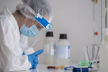 Woman in protective wear sitting at desk examining blood sample - SNF00046