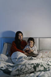 Lachende Mutter und ihre kleine Tochter sitzen zusammen auf dem Bett und schauen sich ein Bilderbuch an - EGAF00046