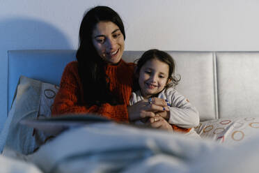 Porträt einer Mutter und ihrer kleinen Tochter, die zusammen auf dem Bett sitzen und ein Bilderbuch anschauen - EGAF00047