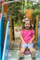 Porträt eines glücklichen kleinen Mädchens, das auf einer Spielplatzrutsche sitzt - MGIF00932