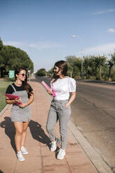 Full length of smiling young female students holding files while walking on footpath at university campus against sky - GRCF00186