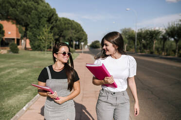 Happy female university students talking while walking on footpath at campus on sunny day - GRCF00187