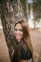 Side view portrait of smiling young woman standing by tree trunk in forest - GRCF00189