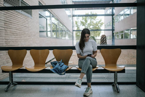 Junge Studentin in voller Länge, die ein Smartphone benutzt, während sie im Korridor einer Universität sitzt - GRCF00192