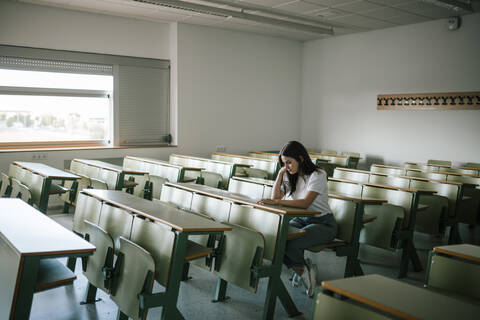 Junge, nachdenkliche Studentin am Schreibtisch sitzend in einem Universitätsklassenzimmer in voller Länge, lizenzfreies Stockfoto