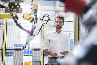 Selbstbewusster Ingenieur betrachtet einen Roboterarm, während er mit einem digitalen Tablet in einer Fabrik steht - DIGF10413