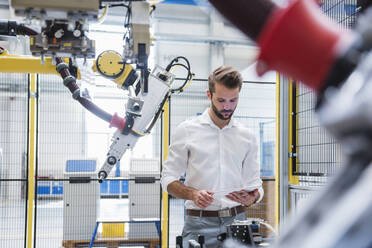 Junger männlicher Experte verwendet ein digitales Tablet, während er an einem Roboterarm in der Industrie steht - DIGF10414