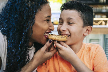 Close-up of cheerful mother and son sharing cookie while sitting at restaurant - DGOF00954