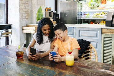Fröhliche Mutter und Sohn benutzen Handys am Tisch im Restaurant - DGOF00961