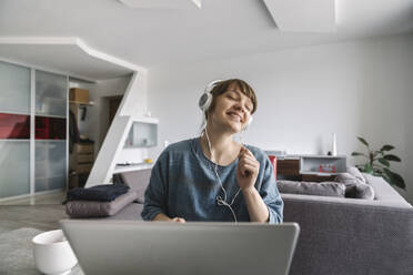 Happy woman wearing headphones and using laptop at home - AHSF02531