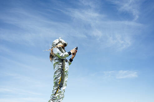 Low angle view of girl in space suit using smart phone while standing against blue sky - JCMF00706