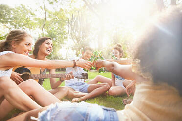 Begeisterte junge Freunde spielen Gitarre und stoßen mit Bierflaschen im sonnigen Sommerpark an - CAIF27212