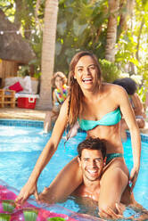 Portrait smiling, confident young couple playing chicken in sunny summer swimming pool - CAIF27216