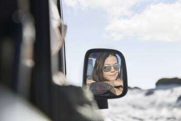 Lächelnde junge Frau mit Sonnenbrille im Seitenspiegel eines Autos - CAIF27243